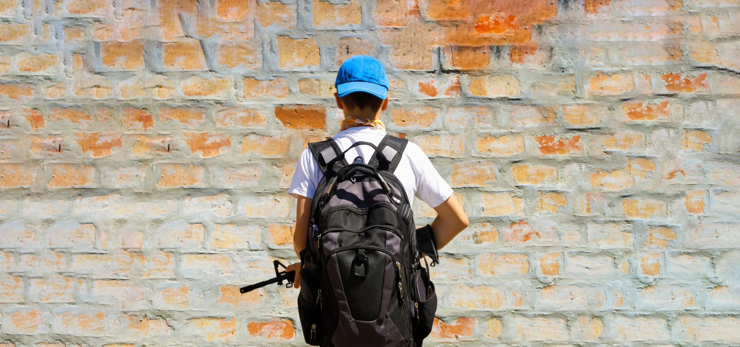 a,young,man,carrying,a,rifle,and,facing,a,wall.